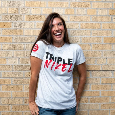 Woman wearing t-shirt with Triple Nikel logo leans against a brick wall